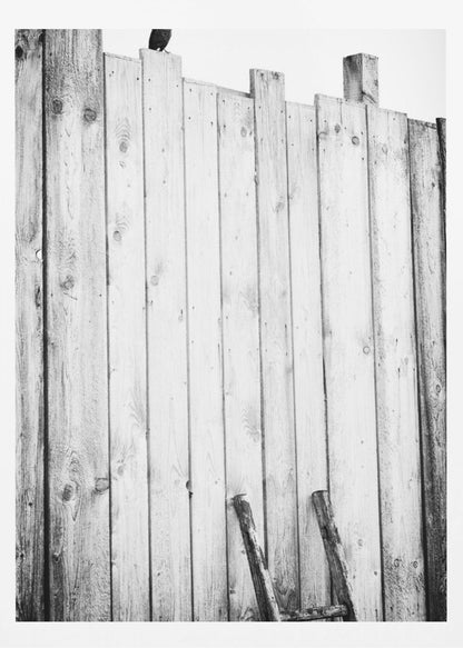 A black and white, low-angle photograph of a weathered wooden plank fence. The vertical planks are of varying heights, creating an uneven top line against a pale sky. The focus is on the texture of the wood, showing prominent grain, knots, and nail holes, all framed in black. Poster