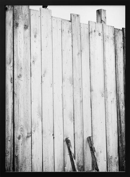 A black and white, low-angle photograph of a weathered wooden plank fence. The vertical planks are of varying heights, creating an uneven top line against a pale sky. The focus is on the texture of the wood, showing prominent grain, knots, and nail holes, all framed in black. Poster