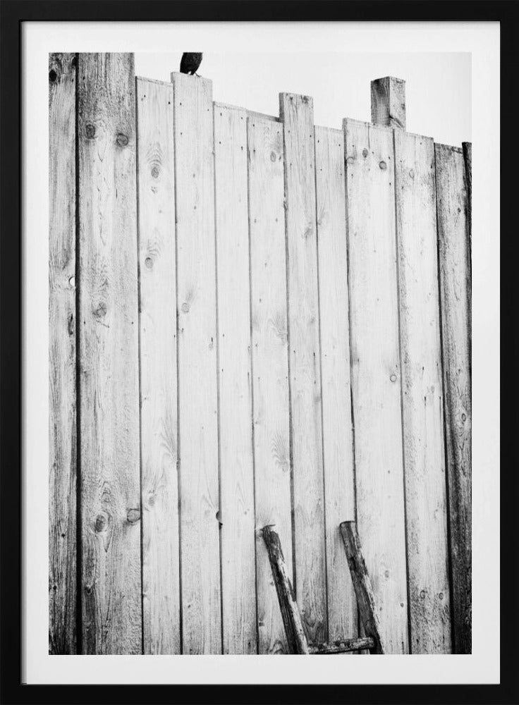 A black and white, low-angle photograph of a weathered wooden plank fence. The vertical planks are of varying heights, creating an uneven top line against a pale sky. The focus is on the texture of the wood, showing prominent grain, knots, and nail holes, all framed in black. Poster