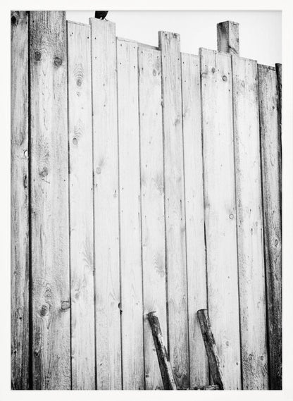 A black and white, low-angle photograph of a weathered wooden plank fence. The vertical planks are of varying heights, creating an uneven top line against a pale sky. The focus is on the texture of the wood, showing prominent grain, knots, and nail holes, all framed in black. Poster