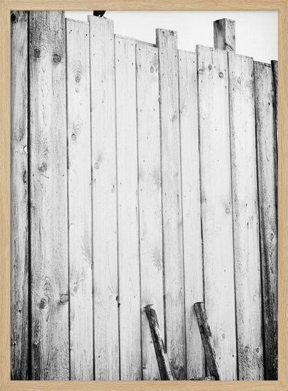 A black and white, low-angle photograph of a weathered wooden plank fence. The vertical planks are of varying heights, creating an uneven top line against a pale sky. The focus is on the texture of the wood, showing prominent grain, knots, and nail holes, all framed in black. Poster