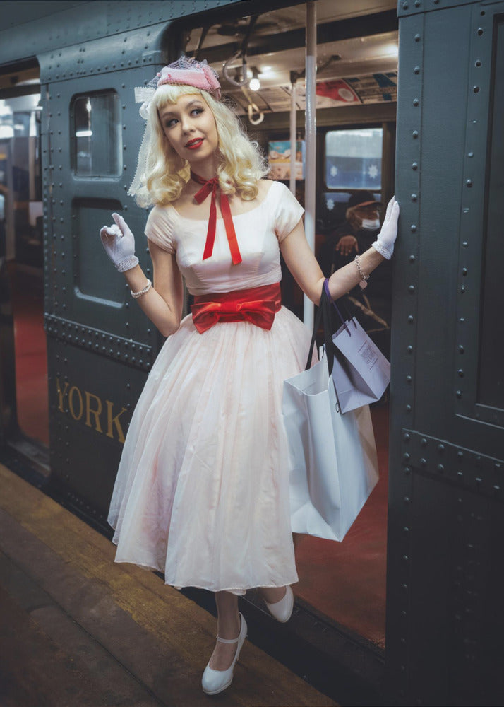 A full-length portrait of a young woman with blonde hair dressed in 1950s-style fashion, including a light pink dress with a bright red sash, a pink veiled hat, and white gloves. She smiles as she steps out of a dark, vintage train car onto a platform, holding shopping bags. Poster