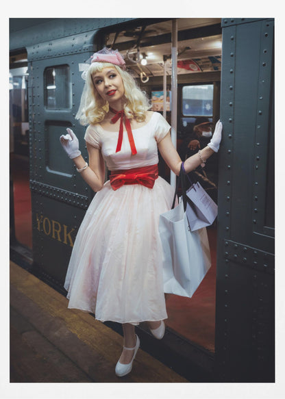 A full-length portrait of a young woman with blonde hair dressed in 1950s-style fashion, including a light pink dress with a bright red sash, a pink veiled hat, and white gloves. She smiles as she steps out of a dark, vintage train car onto a platform, holding shopping bags. Poster