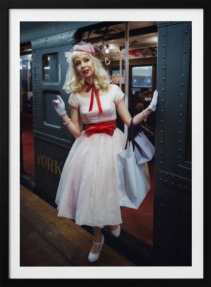 A full-length portrait of a young woman with blonde hair dressed in 1950s-style fashion, including a light pink dress with a bright red sash, a pink veiled hat, and white gloves. She smiles as she steps out of a dark, vintage train car onto a platform, holding shopping bags. Poster