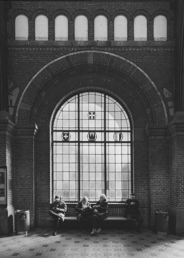 A dramatic black and white photograph of a large, brick-walled waiting area, possibly a train station. Four people are seated on a long bench beneath a massive arched window, which dominates the scene. The lighting creates a moody, contemplative atmosphere, highlighting the grandeur of the architecture. Artwork