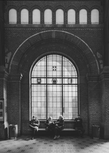 A dramatic black and white photograph of a large, brick-walled waiting area, possibly a train station. Four people are seated on a long bench beneath a massive arched window, which dominates the scene. The lighting creates a moody, contemplative atmosphere, highlighting the grandeur of the architecture. Artwork