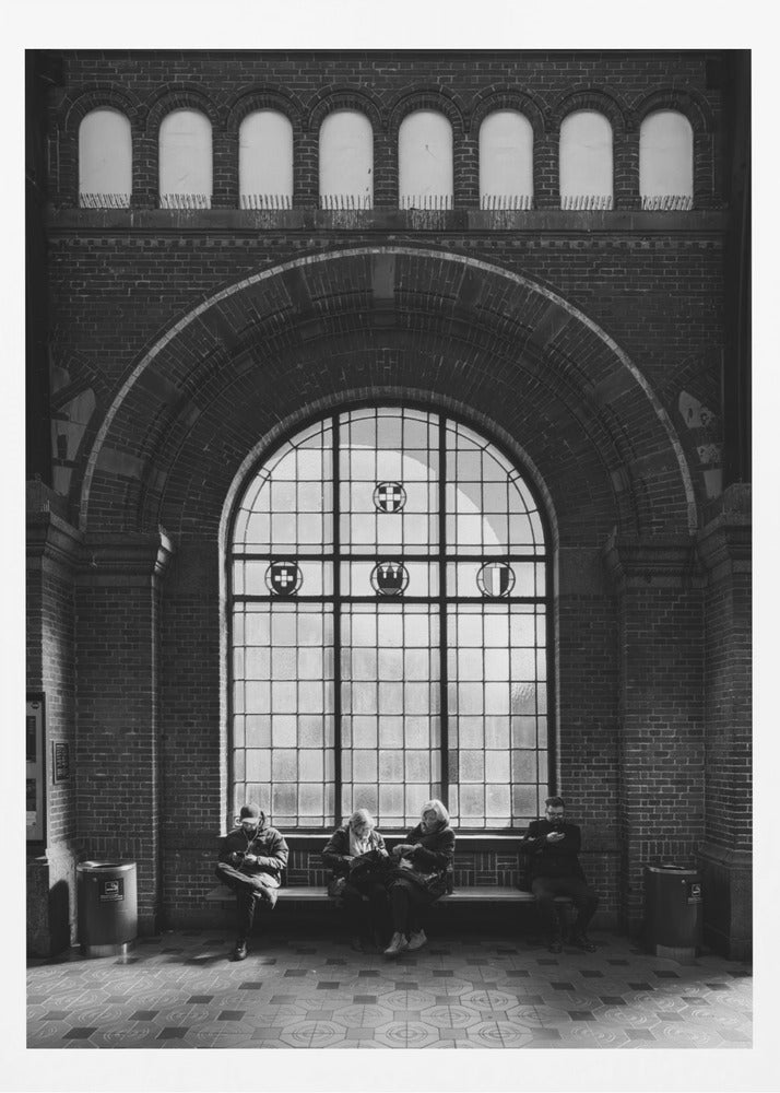 A dramatic black and white photograph of a large, brick-walled waiting area, possibly a train station. Four people are seated on a long bench beneath a massive arched window, which dominates the scene. The lighting creates a moody, contemplative atmosphere, highlighting the grandeur of the architecture. Artwork