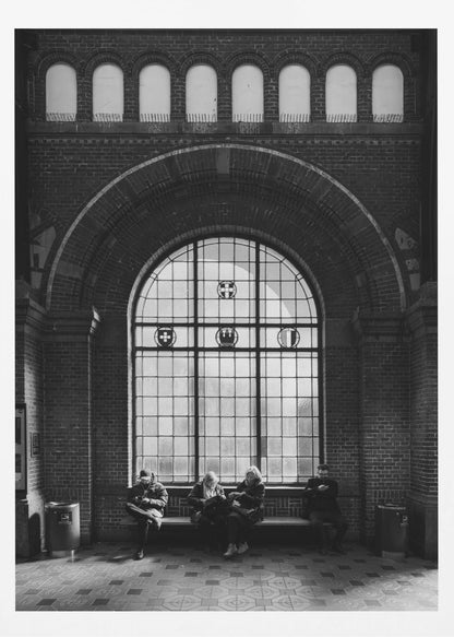 A dramatic black and white photograph of a large, brick-walled waiting area, possibly a train station. Four people are seated on a long bench beneath a massive arched window, which dominates the scene. The lighting creates a moody, contemplative atmosphere, highlighting the grandeur of the architecture. Artwork