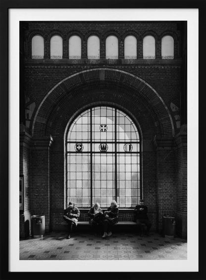 A dramatic black and white photograph of a large, brick-walled waiting area, possibly a train station. Four people are seated on a long bench beneath a massive arched window, which dominates the scene. The lighting creates a moody, contemplative atmosphere, highlighting the grandeur of the architecture. Artwork