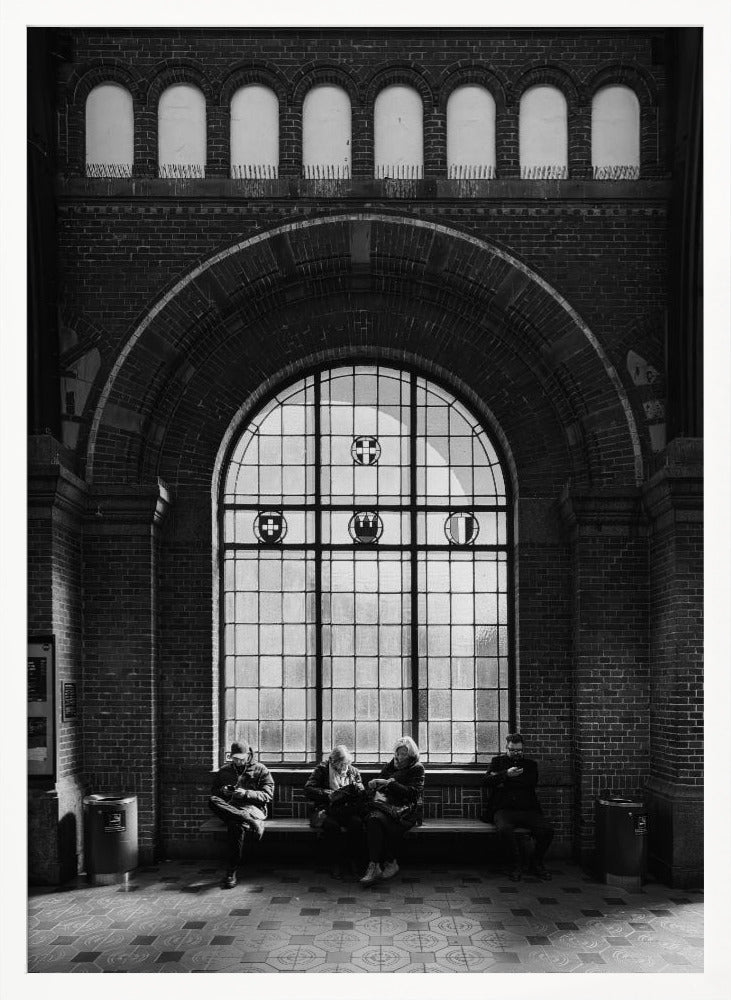 A dramatic black and white photograph of a large, brick-walled waiting area, possibly a train station. Four people are seated on a long bench beneath a massive arched window, which dominates the scene. The lighting creates a moody, contemplative atmosphere, highlighting the grandeur of the architecture. Artwork