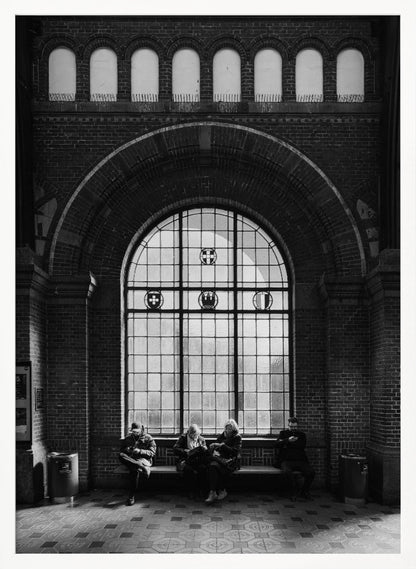 A dramatic black and white photograph of a large, brick-walled waiting area, possibly a train station. Four people are seated on a long bench beneath a massive arched window, which dominates the scene. The lighting creates a moody, contemplative atmosphere, highlighting the grandeur of the architecture. Artwork