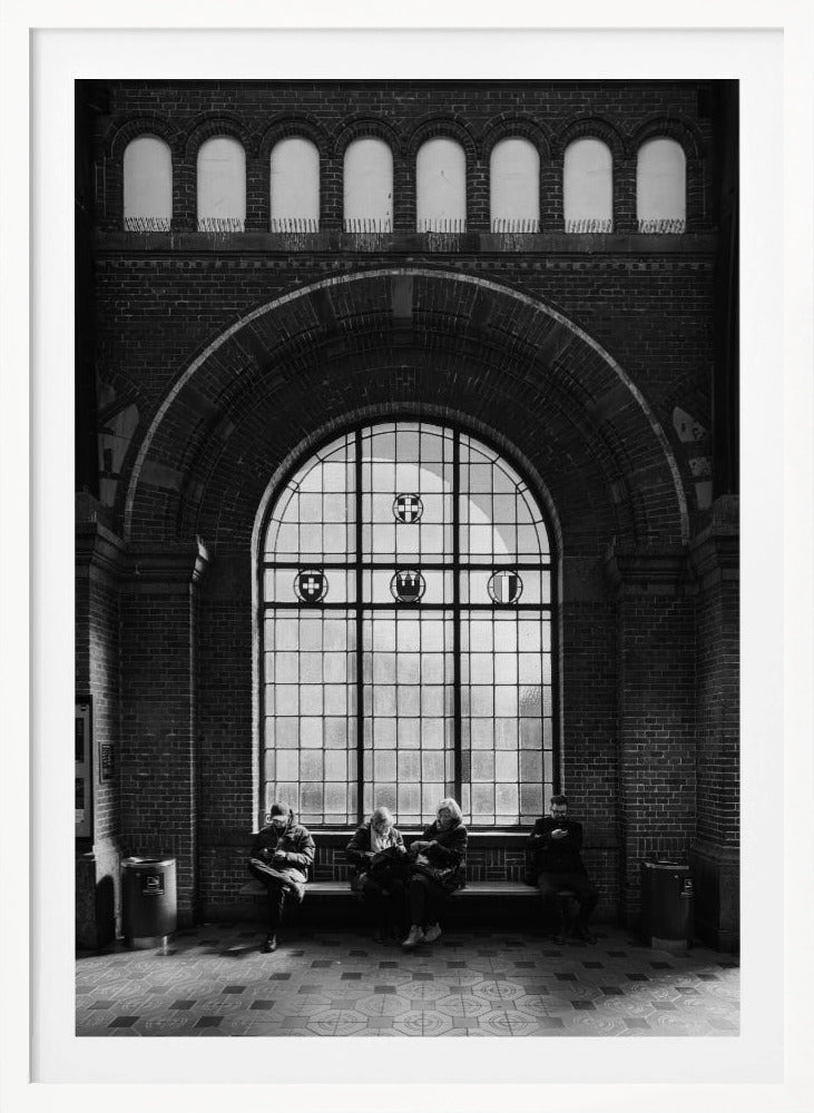 A dramatic black and white photograph of a large, brick-walled waiting area, possibly a train station. Four people are seated on a long bench beneath a massive arched window, which dominates the scene. The lighting creates a moody, contemplative atmosphere, highlighting the grandeur of the architecture. Artwork