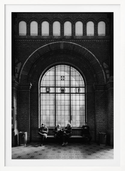 A dramatic black and white photograph of a large, brick-walled waiting area, possibly a train station. Four people are seated on a long bench beneath a massive arched window, which dominates the scene. The lighting creates a moody, contemplative atmosphere, highlighting the grandeur of the architecture. Artwork