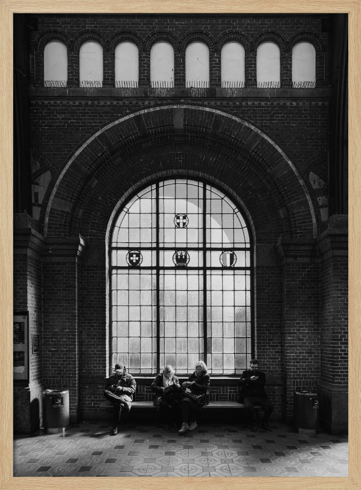 A dramatic black and white photograph of a large, brick-walled waiting area, possibly a train station. Four people are seated on a long bench beneath a massive arched window, which dominates the scene. The lighting creates a moody, contemplative atmosphere, highlighting the grandeur of the architecture. Artwork