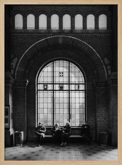 A dramatic black and white photograph of a large, brick-walled waiting area, possibly a train station. Four people are seated on a long bench beneath a massive arched window, which dominates the scene. The lighting creates a moody, contemplative atmosphere, highlighting the grandeur of the architecture. Artwork