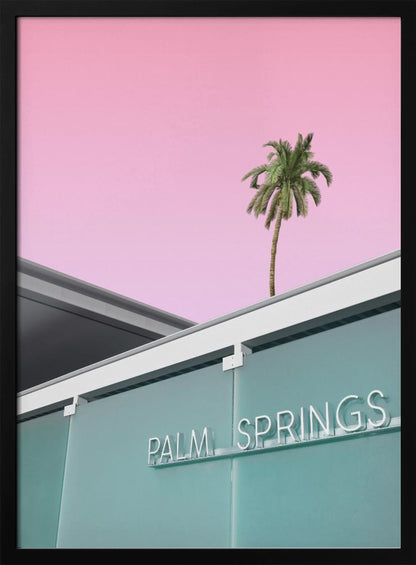 A low-angle shot of a modern building with a frosted teal glass facade and a white neon sign that reads 'PALM SPRINGS'. Above the building, a single tall palm tree stands against a solid, vibrant pink sky, creating a minimalist and stylized composition. Decor