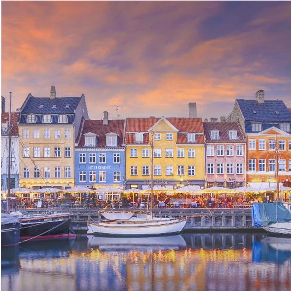 A framed photograph of the Nyhavn harbor in Copenhagen at dusk, featuring a row of brightly colored historic buildings along the canal. Sailboats are moored in the water, which reflects the warm glow of the building lights and the vibrant orange and purple sunset sky. Print