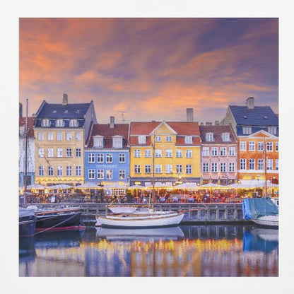 A framed photograph of the Nyhavn harbor in Copenhagen at dusk, featuring a row of brightly colored historic buildings along the canal. Sailboats are moored in the water, which reflects the warm glow of the building lights and the vibrant orange and purple sunset sky. Print