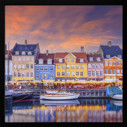 A framed photograph of the Nyhavn harbor in Copenhagen at dusk, featuring a row of brightly colored historic buildings along the canal. Sailboats are moored in the water, which reflects the warm glow of the building lights and the vibrant orange and purple sunset sky. Print