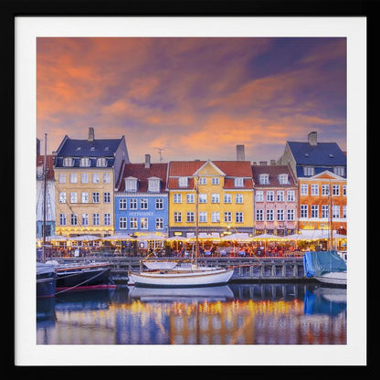 A framed photograph of the Nyhavn harbor in Copenhagen at dusk, featuring a row of brightly colored historic buildings along the canal. Sailboats are moored in the water, which reflects the warm glow of the building lights and the vibrant orange and purple sunset sky. Print