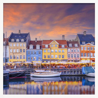 A framed photograph of the Nyhavn harbor in Copenhagen at dusk, featuring a row of brightly colored historic buildings along the canal. Sailboats are moored in the water, which reflects the warm glow of the building lights and the vibrant orange and purple sunset sky. Print