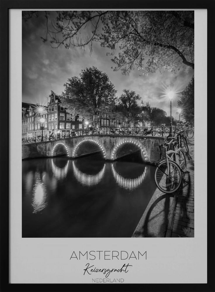 A black and white poster of Amsterdam's Keizersgracht canal at night. An arched bridge with glowing lights is the centerpiece, its reflection shimmering on the dark water. Bicycles are parked along the bridge and in the foreground on a cobblestone path. Classic canal houses and trees line the background under a dramatic, cloudy sky. The image is framed with text below reading 'AMSTERDAM Keizersgracht NEDERLAND'. Artwork