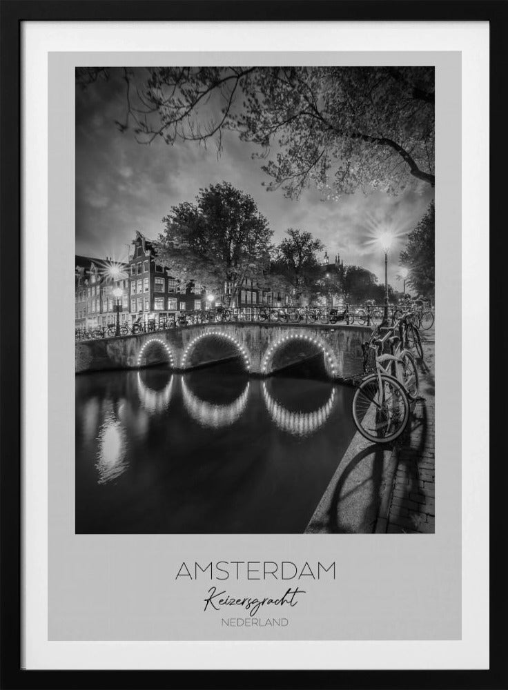 A black and white poster of Amsterdam's Keizersgracht canal at night. An arched bridge with glowing lights is the centerpiece, its reflection shimmering on the dark water. Bicycles are parked along the bridge and in the foreground on a cobblestone path. Classic canal houses and trees line the background under a dramatic, cloudy sky. The image is framed with text below reading 'AMSTERDAM Keizersgracht NEDERLAND'. Artwork