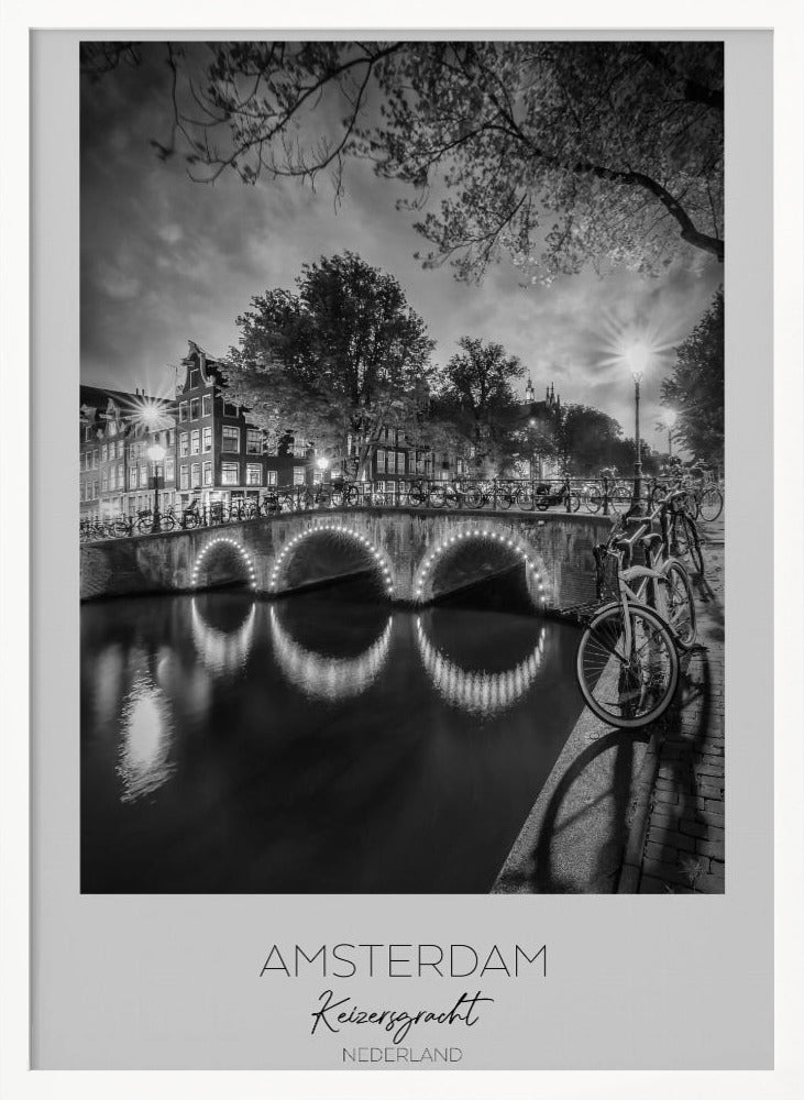 A black and white poster of Amsterdam's Keizersgracht canal at night. An arched bridge with glowing lights is the centerpiece, its reflection shimmering on the dark water. Bicycles are parked along the bridge and in the foreground on a cobblestone path. Classic canal houses and trees line the background under a dramatic, cloudy sky. The image is framed with text below reading 'AMSTERDAM Keizersgracht NEDERLAND'. Artwork