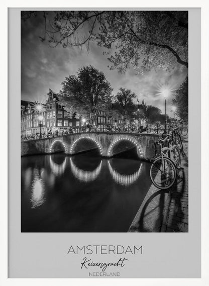 A black and white poster of Amsterdam's Keizersgracht canal at night. An arched bridge with glowing lights is the centerpiece, its reflection shimmering on the dark water. Bicycles are parked along the bridge and in the foreground on a cobblestone path. Classic canal houses and trees line the background under a dramatic, cloudy sky. The image is framed with text below reading 'AMSTERDAM Keizersgracht NEDERLAND'. Artwork