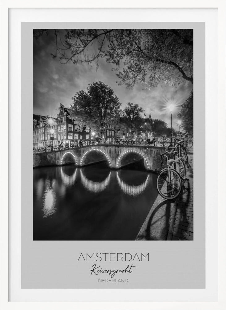 A black and white poster of Amsterdam's Keizersgracht canal at night. An arched bridge with glowing lights is the centerpiece, its reflection shimmering on the dark water. Bicycles are parked along the bridge and in the foreground on a cobblestone path. Classic canal houses and trees line the background under a dramatic, cloudy sky. The image is framed with text below reading 'AMSTERDAM Keizersgracht NEDERLAND'. Artwork