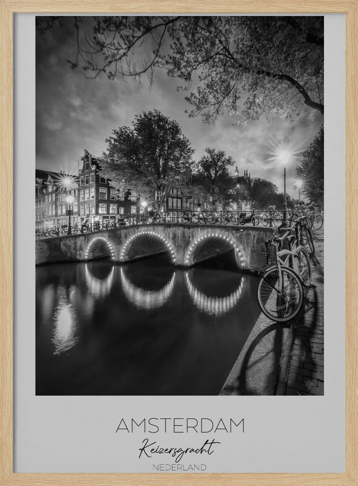 A black and white poster of Amsterdam's Keizersgracht canal at night. An arched bridge with glowing lights is the centerpiece, its reflection shimmering on the dark water. Bicycles are parked along the bridge and in the foreground on a cobblestone path. Classic canal houses and trees line the background under a dramatic, cloudy sky. The image is framed with text below reading 'AMSTERDAM Keizersgracht NEDERLAND'. Artwork