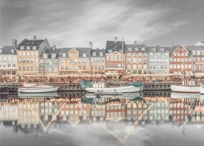 A framed, color-selective photograph of the Nyhavn harbor in Copenhagen. A row of colorful 17th and 18th-century townhouses lines the waterfront, with their warm lights and facades reflecting in the calm canal. The sky and parts of the water are in black and white, making the colored buildings stand out. Several boats are moored along the quay. Decor