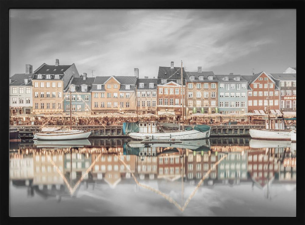 A framed, color-selective photograph of the Nyhavn harbor in Copenhagen. A row of colorful 17th and 18th-century townhouses lines the waterfront, with their warm lights and facades reflecting in the calm canal. The sky and parts of the water are in black and white, making the colored buildings stand out. Several boats are moored along the quay. Decor