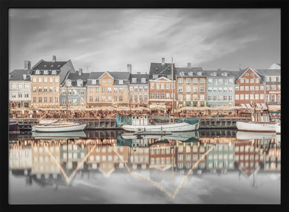 A framed, color-selective photograph of the Nyhavn harbor in Copenhagen. A row of colorful 17th and 18th-century townhouses lines the waterfront, with their warm lights and facades reflecting in the calm canal. The sky and parts of the water are in black and white, making the colored buildings stand out. Several boats are moored along the quay. Decor