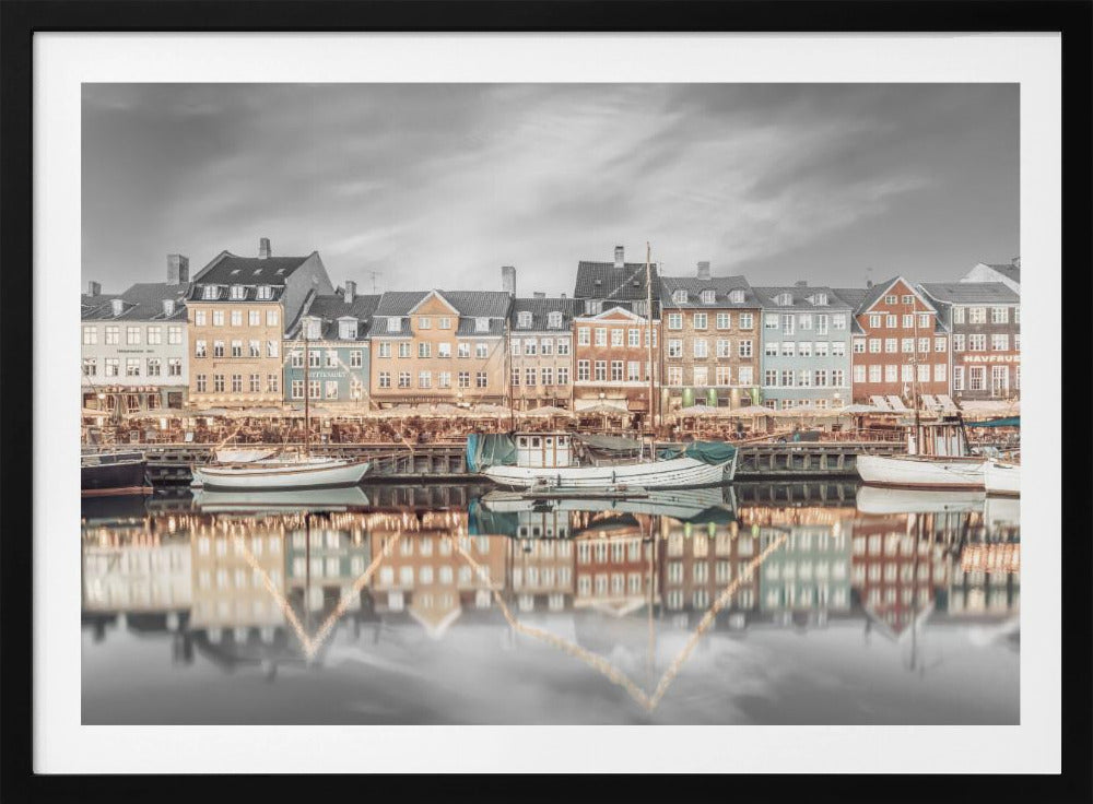 A framed, color-selective photograph of the Nyhavn harbor in Copenhagen. A row of colorful 17th and 18th-century townhouses lines the waterfront, with their warm lights and facades reflecting in the calm canal. The sky and parts of the water are in black and white, making the colored buildings stand out. Several boats are moored along the quay. Decor