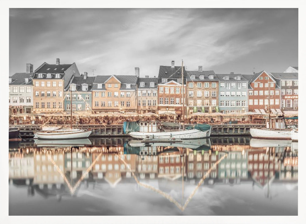 A framed, color-selective photograph of the Nyhavn harbor in Copenhagen. A row of colorful 17th and 18th-century townhouses lines the waterfront, with their warm lights and facades reflecting in the calm canal. The sky and parts of the water are in black and white, making the colored buildings stand out. Several boats are moored along the quay. Decor