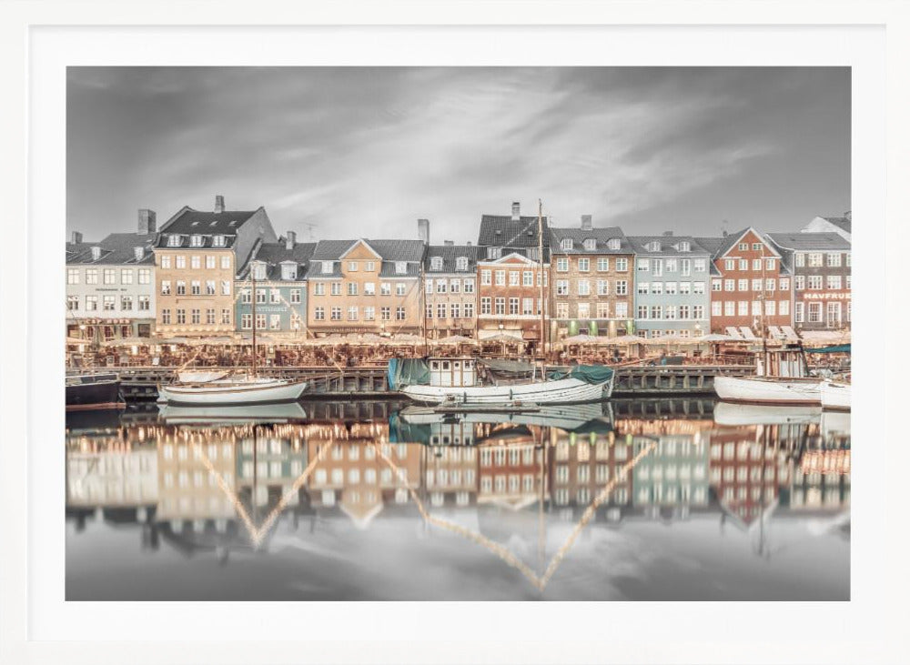 A framed, color-selective photograph of the Nyhavn harbor in Copenhagen. A row of colorful 17th and 18th-century townhouses lines the waterfront, with their warm lights and facades reflecting in the calm canal. The sky and parts of the water are in black and white, making the colored buildings stand out. Several boats are moored along the quay. Decor