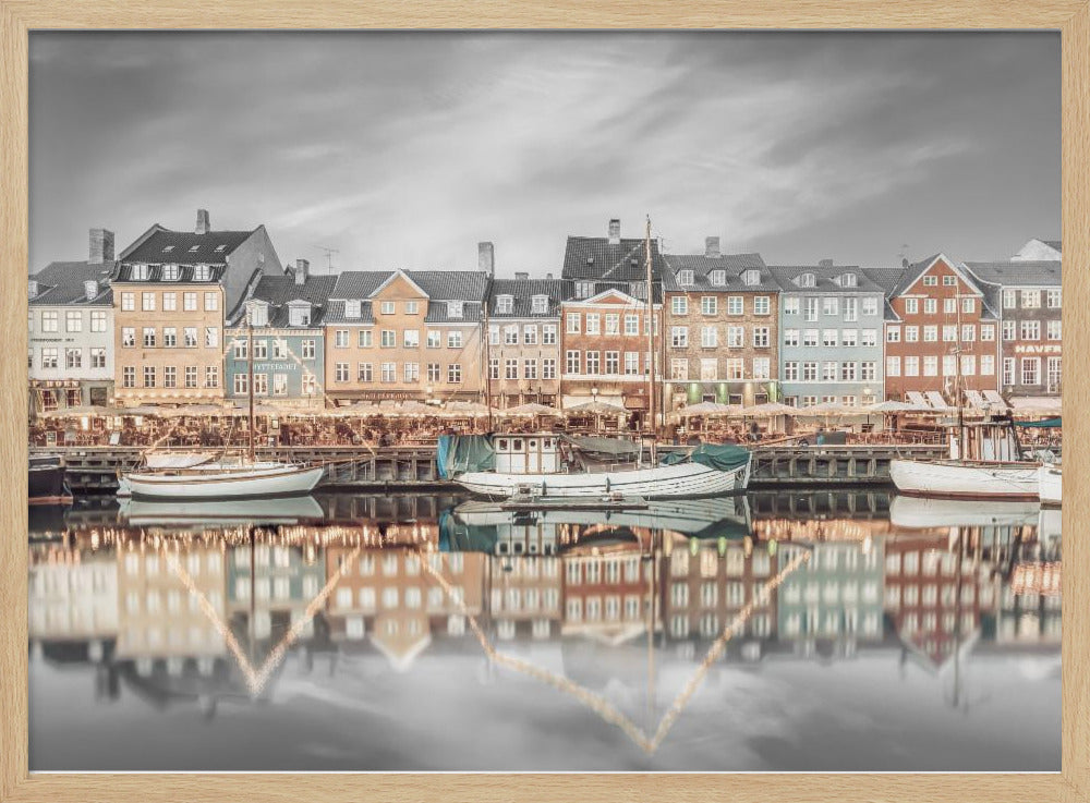 A framed, color-selective photograph of the Nyhavn harbor in Copenhagen. A row of colorful 17th and 18th-century townhouses lines the waterfront, with their warm lights and facades reflecting in the calm canal. The sky and parts of the water are in black and white, making the colored buildings stand out. Several boats are moored along the quay. Decor