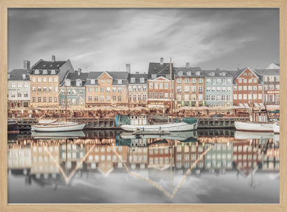 A framed, color-selective photograph of the Nyhavn harbor in Copenhagen. A row of colorful 17th and 18th-century townhouses lines the waterfront, with their warm lights and facades reflecting in the calm canal. The sky and parts of the water are in black and white, making the colored buildings stand out. Several boats are moored along the quay. Decor
