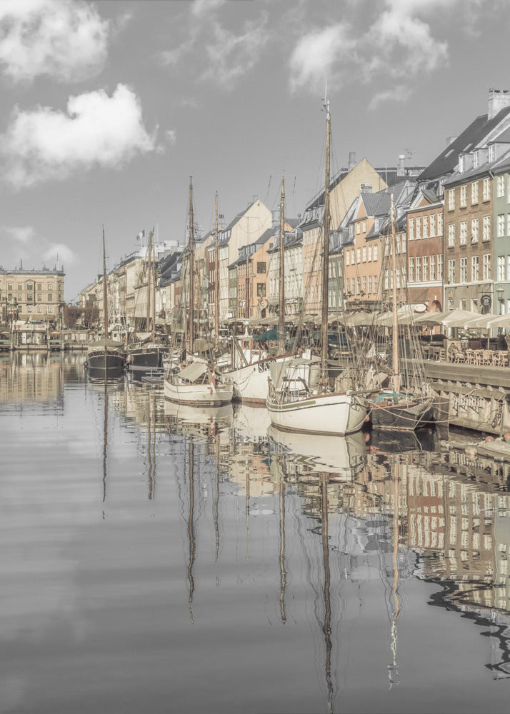 A muted, desaturated photograph of the Nyhavn harbor in Copenhagen. Old-fashioned sailboats are moored along the canal in front of a row of colorful historic buildings. The boats, buildings, and a cloudy sky are perfectly reflected in the calm water. Wall Art