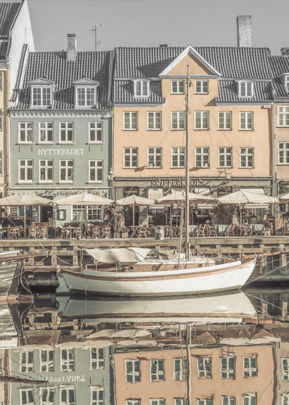 A scenic photograph of a European harbor with colorful buildings reflected in the water. A white sailboat is moored in the foreground in front of a pale green and a yellow gabled building. The image has a selective color effect, with the building facades in muted tones and the sky and roofs in black and white. Artwork
