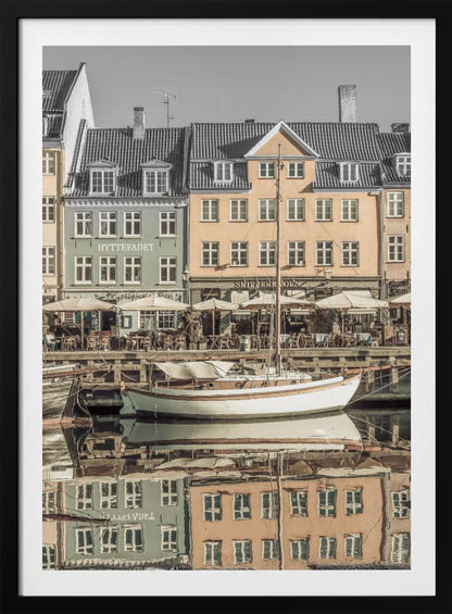 A scenic photograph of a European harbor with colorful buildings reflected in the water. A white sailboat is moored in the foreground in front of a pale green and a yellow gabled building. The image has a selective color effect, with the building facades in muted tones and the sky and roofs in black and white. Artwork