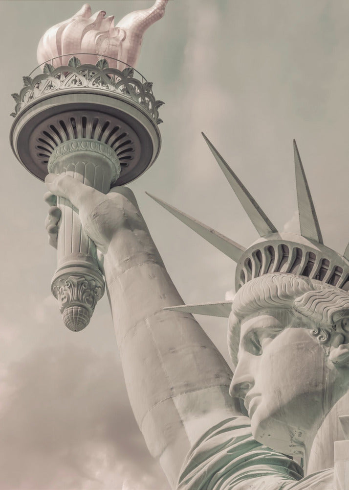 A low-angle, close-up photograph of the Statue of Liberty with a vintage color treatment. The shot captures her profile, crown, and her arm holding the torch aloft against a muted, cloudy sky, all enclosed in a thin black frame. Poster