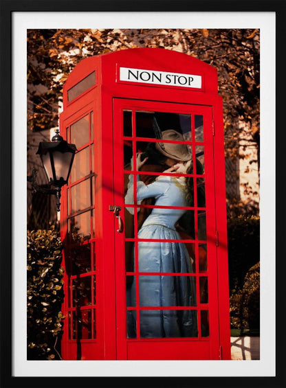 A couple in vintage clothing share a passionate kiss inside a classic bright red telephone booth. The woman is wearing a light blue dress, and the man wears a hat. The sign above the booth door reads 'NON STOP'. The setting appears to be outdoors in autumn. Wall Art