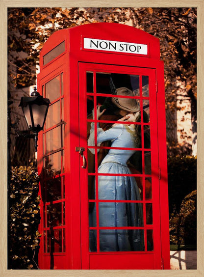A couple in vintage clothing share a passionate kiss inside a classic bright red telephone booth. The woman is wearing a light blue dress, and the man wears a hat. The sign above the booth door reads 'NON STOP'. The setting appears to be outdoors in autumn. Wall Art