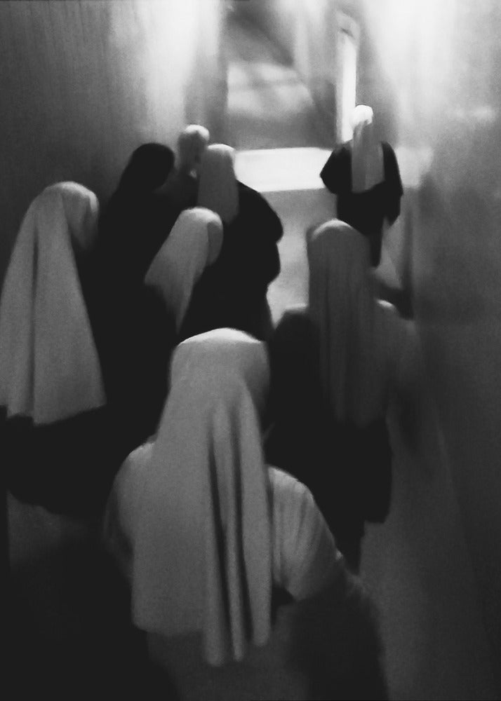 A mysterious black and white photograph captures a group of robed figures, possibly nuns, from above as they walk down a dark, narrow corridor. The image is blurred, creating a ghostly, ethereal atmosphere as the figures' white head coverings stand out against the deep shadows. Artwork