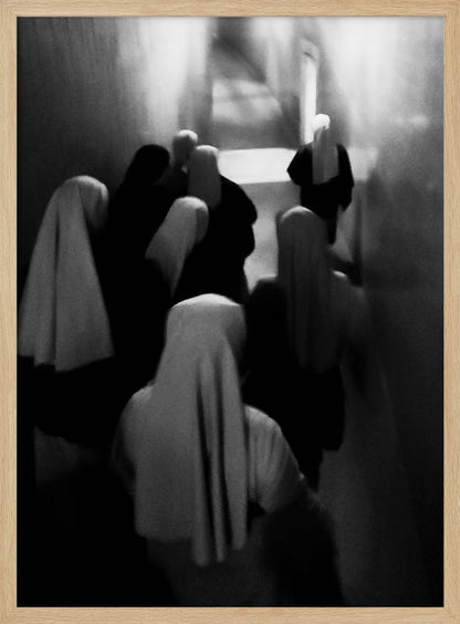 A mysterious black and white photograph captures a group of robed figures, possibly nuns, from above as they walk down a dark, narrow corridor. The image is blurred, creating a ghostly, ethereal atmosphere as the figures' white head coverings stand out against the deep shadows. Artwork