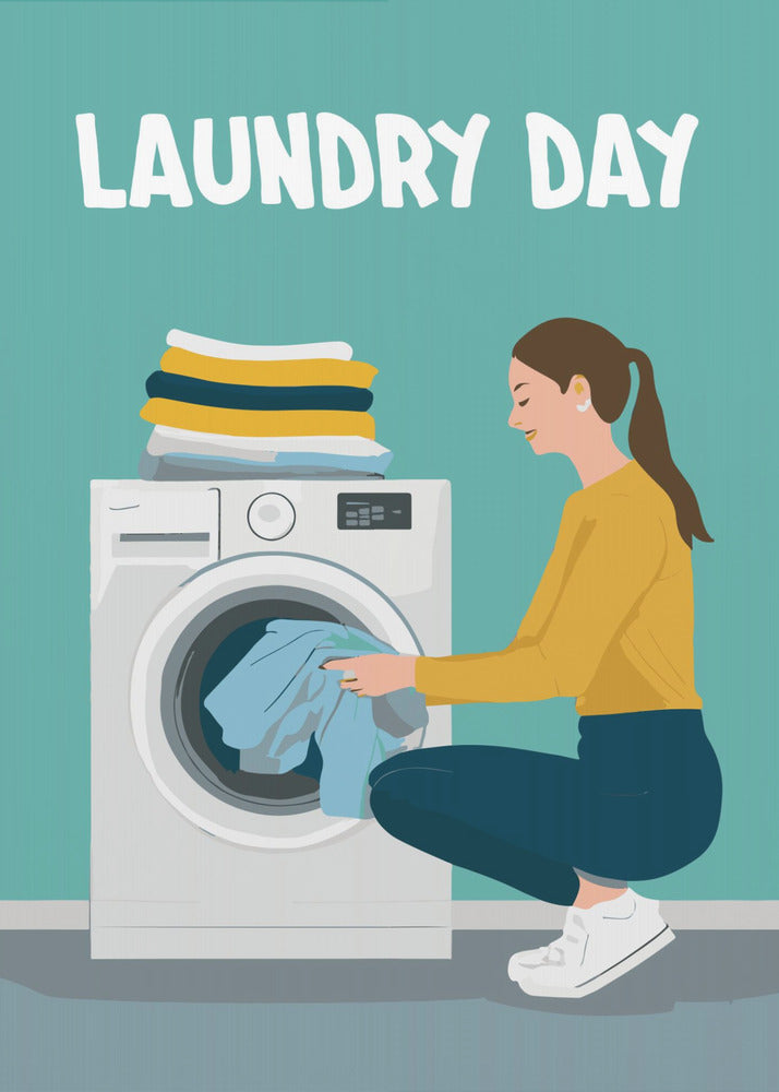 A framed illustration poster with the words 'LAUNDRY DAY' in white at the top. A woman with brown hair in a ponytail, wearing a yellow top and blue pants, crouches to load blue clothes into a white front-loading washing machine. A stack of folded laundry sits atop the machine against a teal background. Print