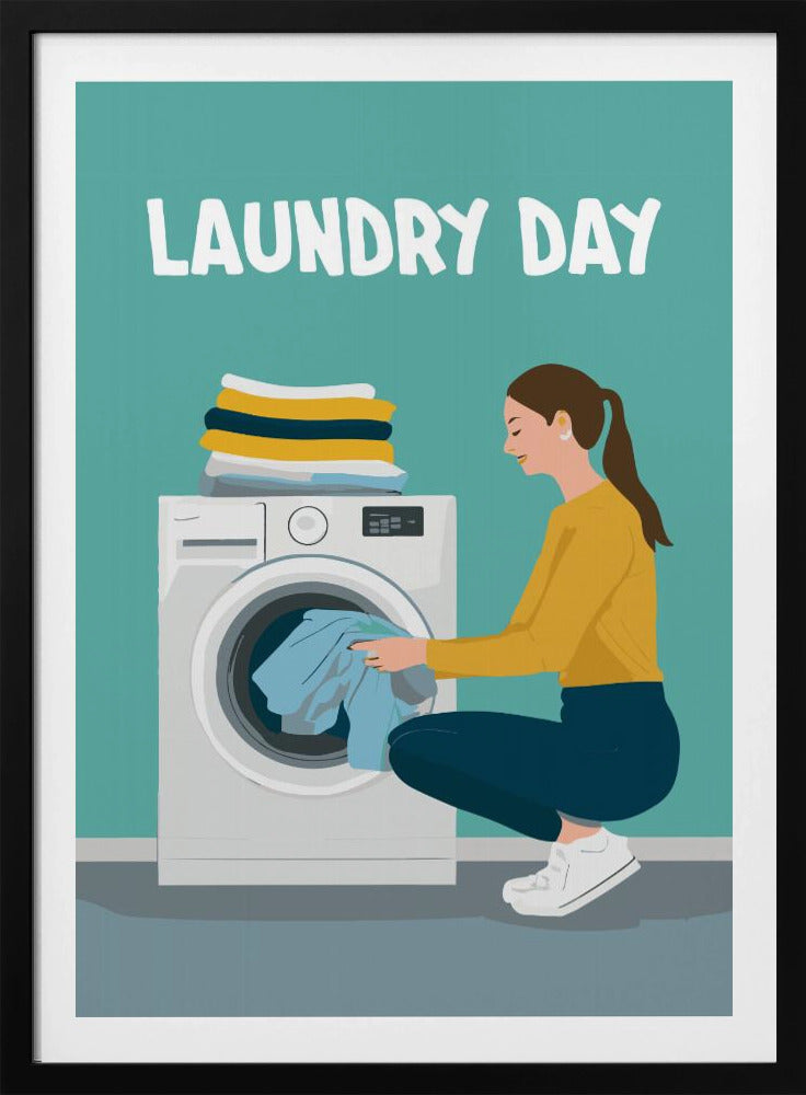 A framed illustration poster with the words 'LAUNDRY DAY' in white at the top. A woman with brown hair in a ponytail, wearing a yellow top and blue pants, crouches to load blue clothes into a white front-loading washing machine. A stack of folded laundry sits atop the machine against a teal background. Print
