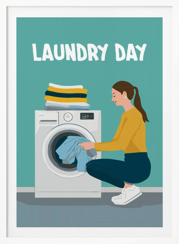 A framed illustration poster with the words 'LAUNDRY DAY' in white at the top. A woman with brown hair in a ponytail, wearing a yellow top and blue pants, crouches to load blue clothes into a white front-loading washing machine. A stack of folded laundry sits atop the machine against a teal background. Print
